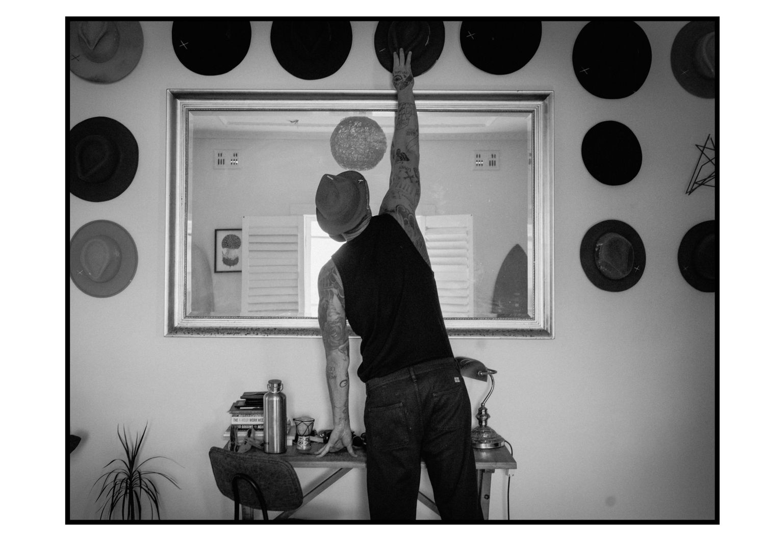 Tattooed man reaching up to arrange hats displayed on white studio wall.