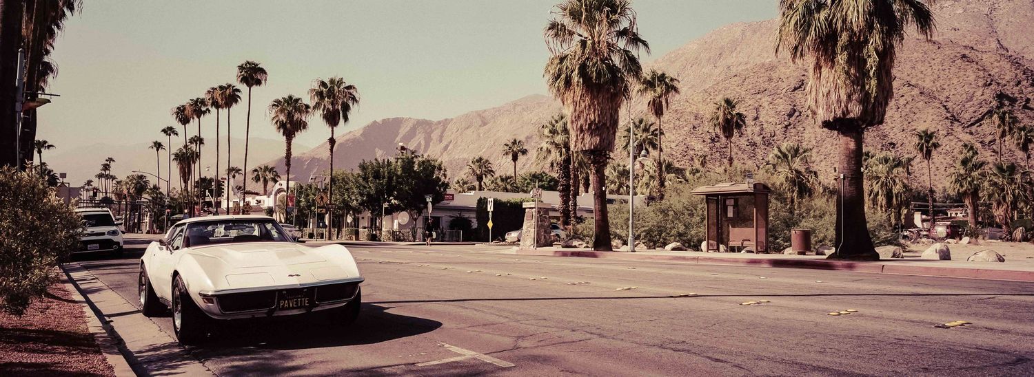 Vintage panoramic street scene in Palm Springs with a classic white Corvette and swaying palm trees.