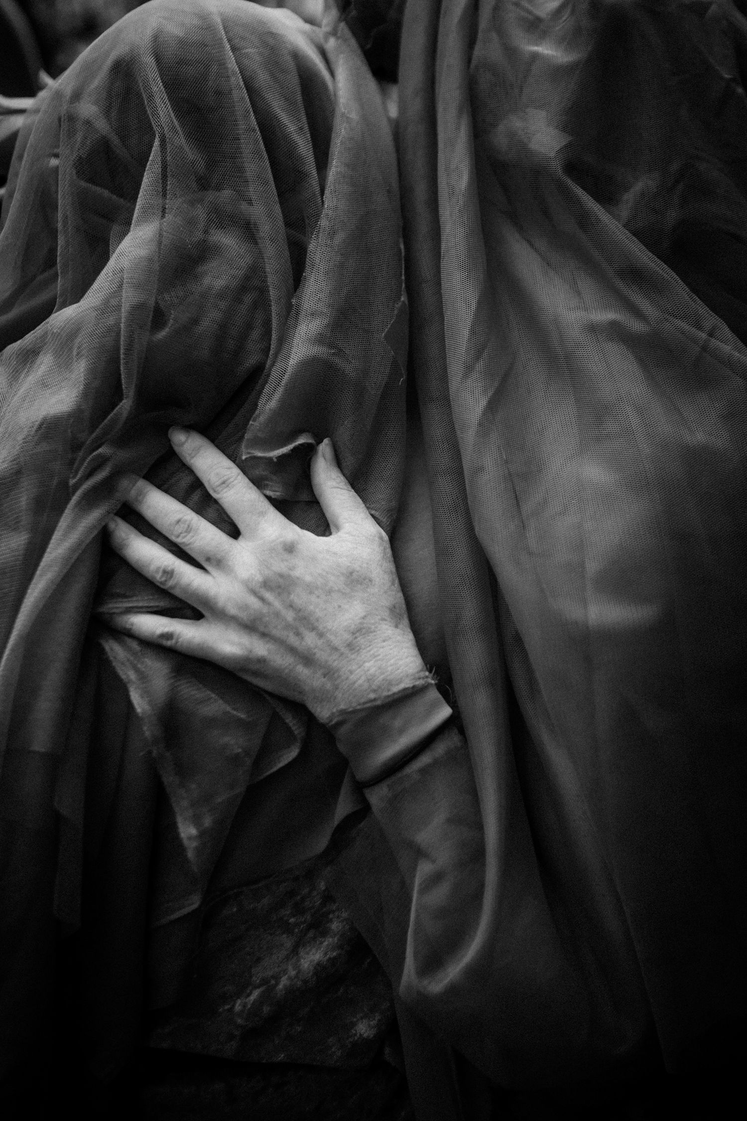 Black and white artistic photo of a figure draped in sheer fabric with a hand on chest.