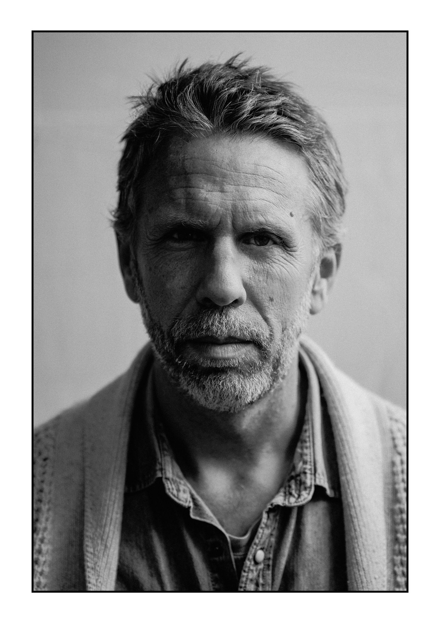 Close-up black and white portrait of weathered bearded man in denim shirt with intense direct gaze.
