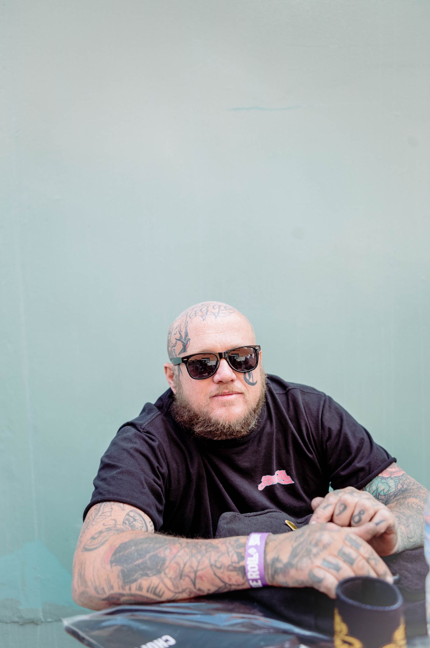 Heavily tattooed man with sunglasses and beard sitting at a table against a mint wall.
