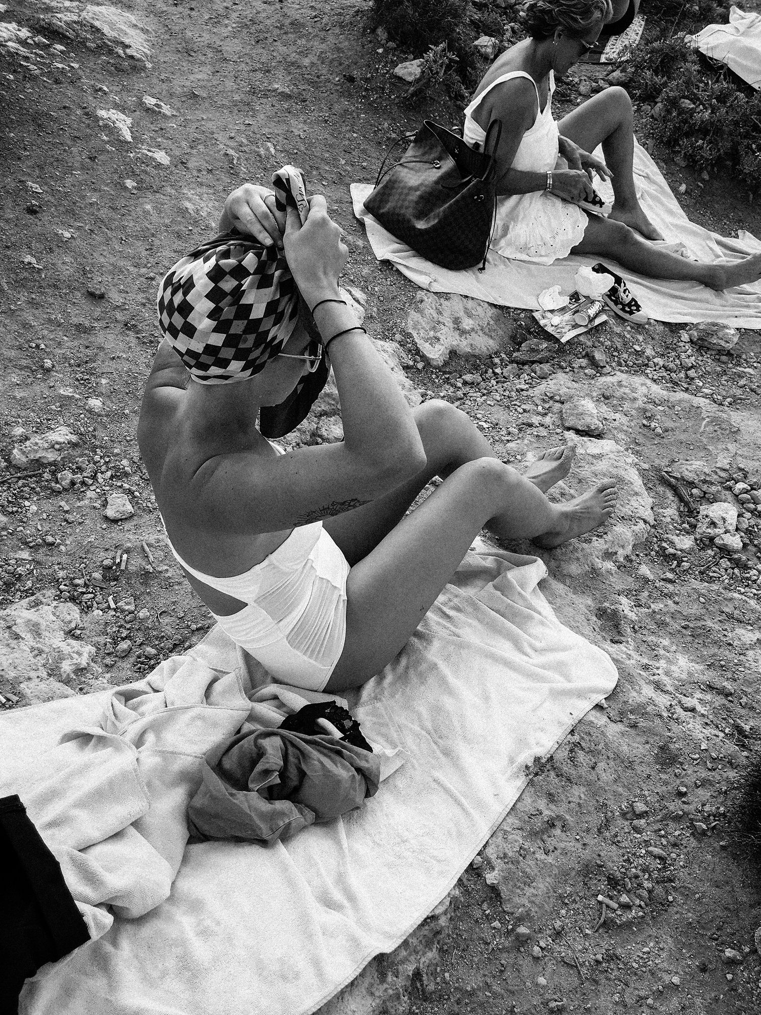Black and white photo of women sunbathing on towels on rocky ground outdoors.