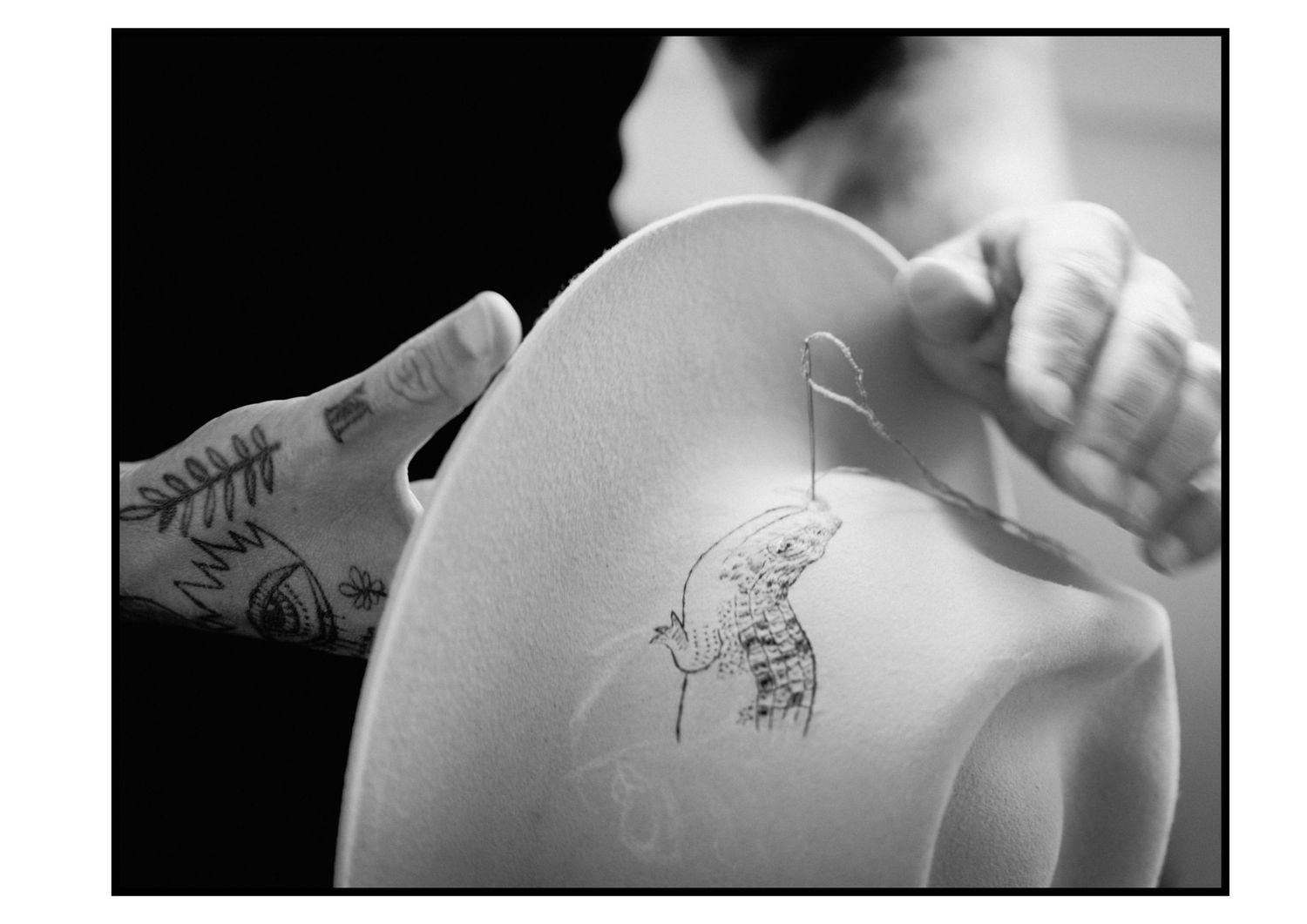 Close-up of tattooed hands holding white felt hat with delicate line drawing.