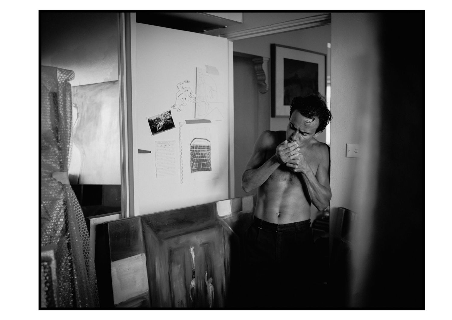 Shirtless athletic man examines something in hands inside art studio with canvases in background.