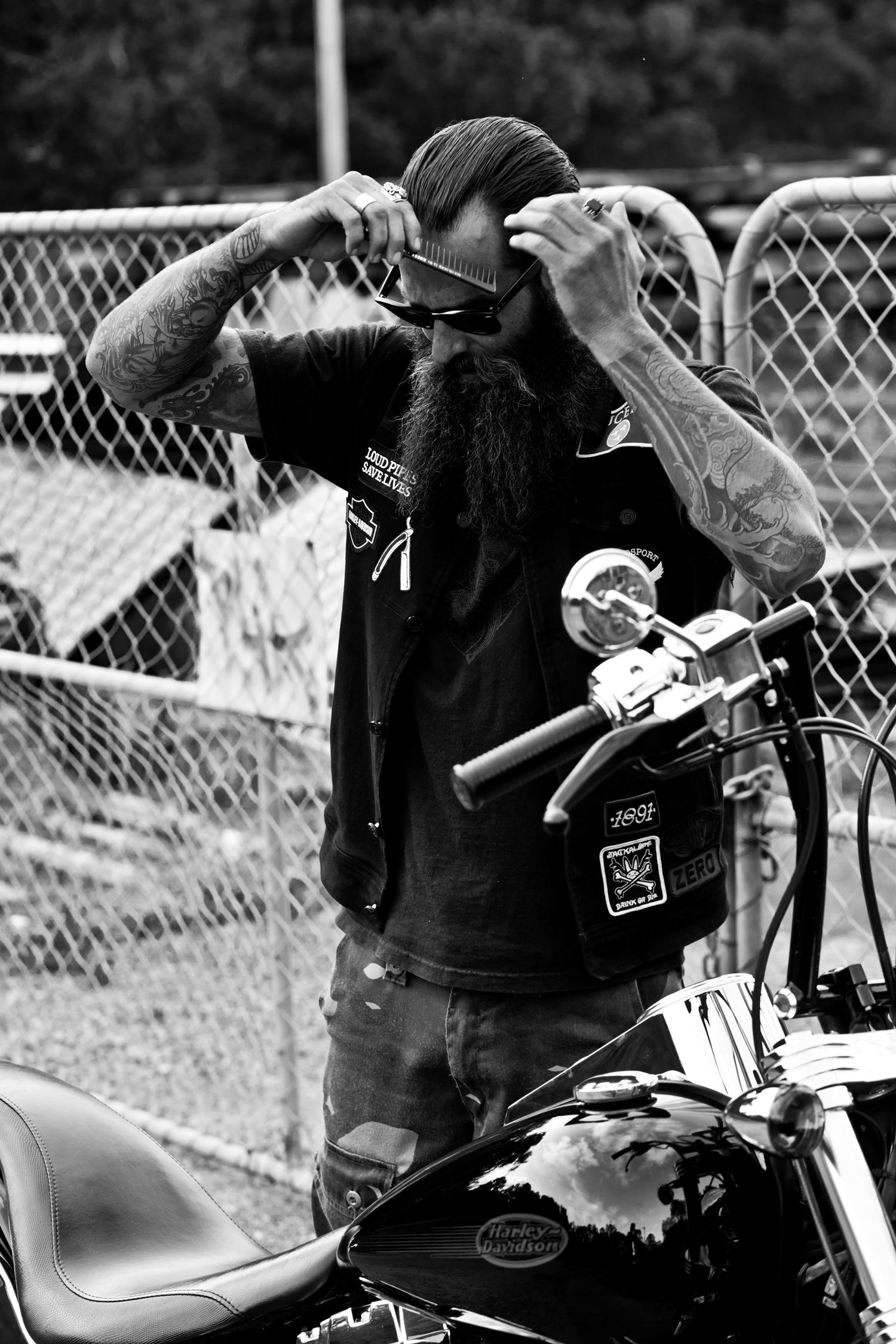 Tattooed bearded biker adjusting sunglasses beside a Harley-Davidson motorcycle in black and white.