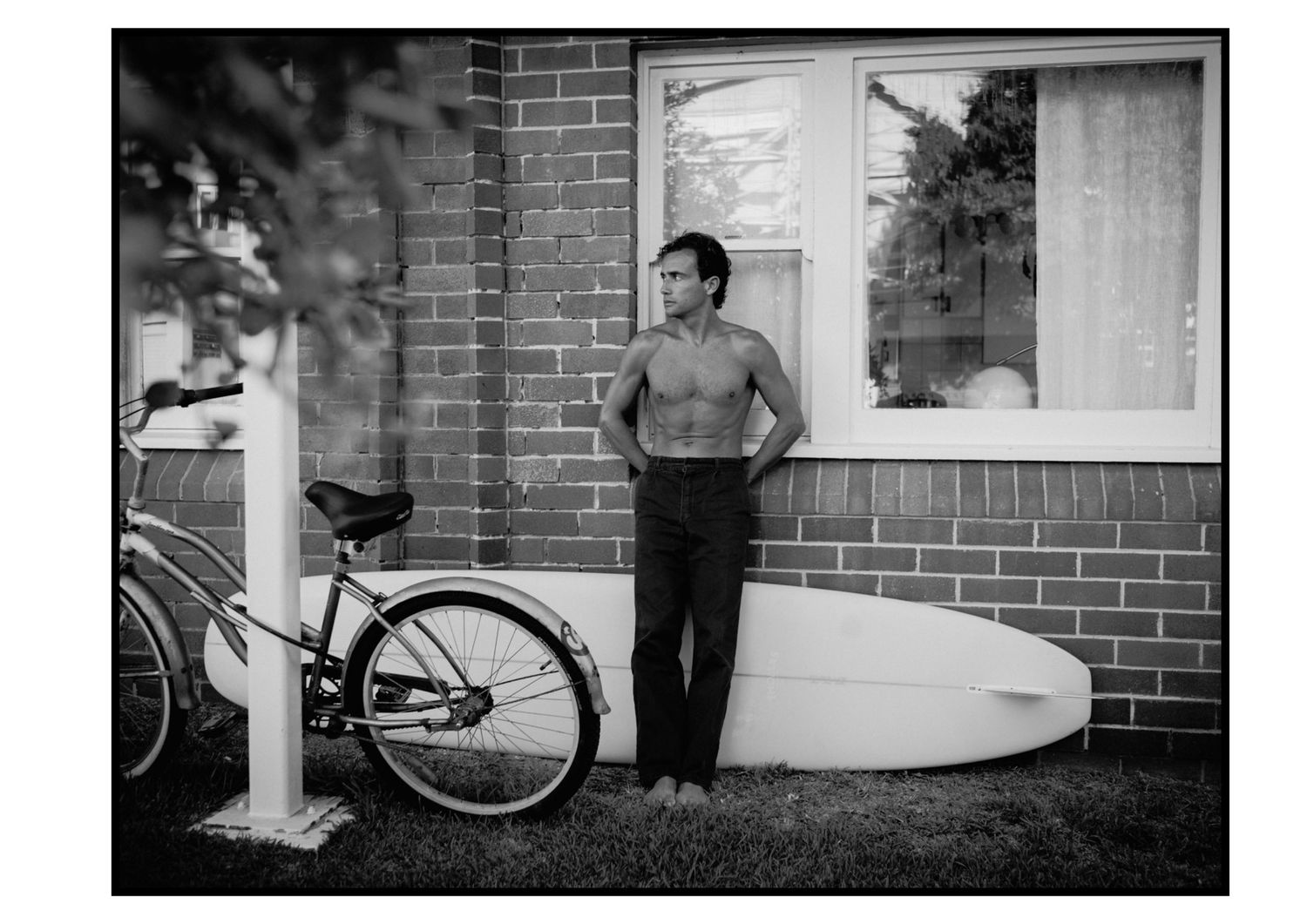 Shirtless man leaning against brick wall with surfboard and bicycle in black and white photo.