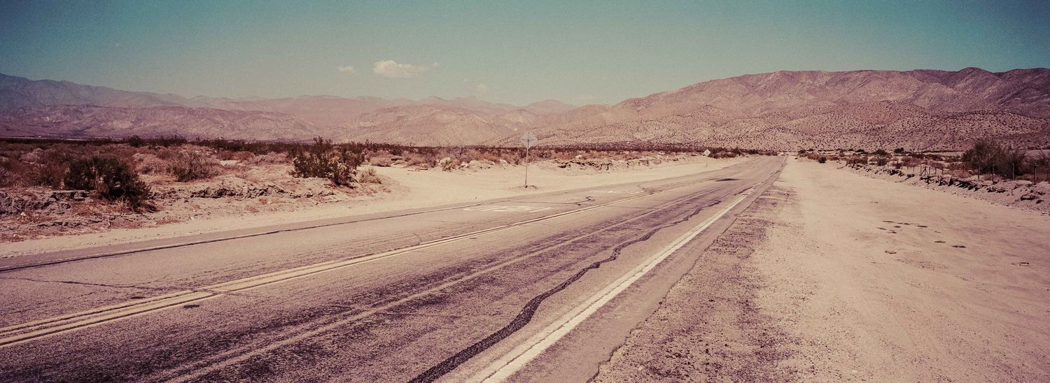 Panoramic view of a long straight desert highway stretching toward distant mountains under a hazy sky.