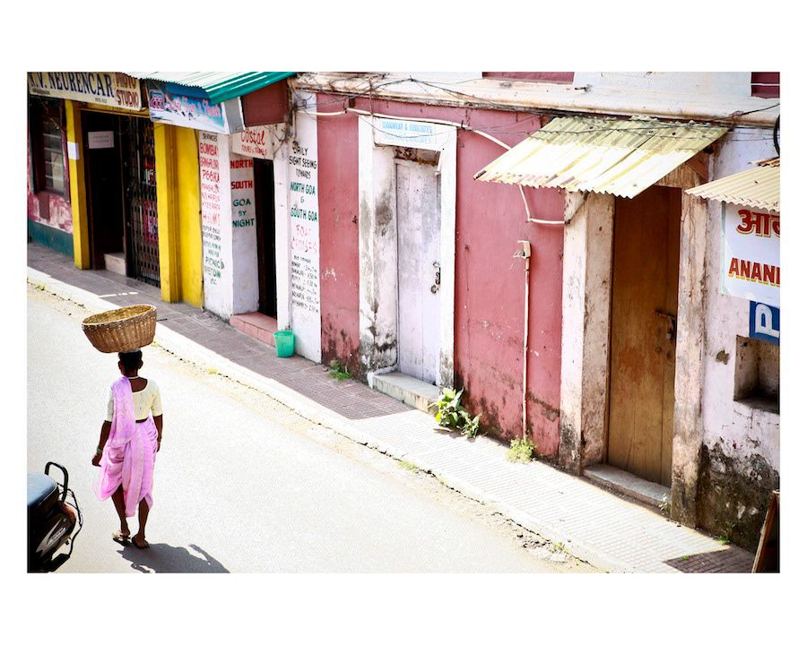 A woman balancing a basket on her head walks along a quiet colorful street in a small Indian town.