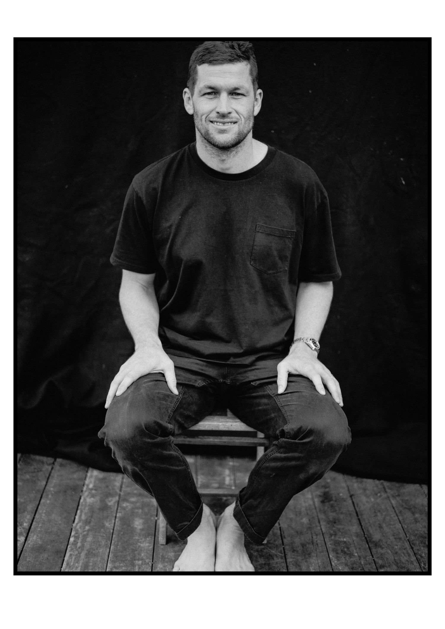 Smiling man in black t-shirt and jeans sits relaxed on a stool against dark background in black and white photo.