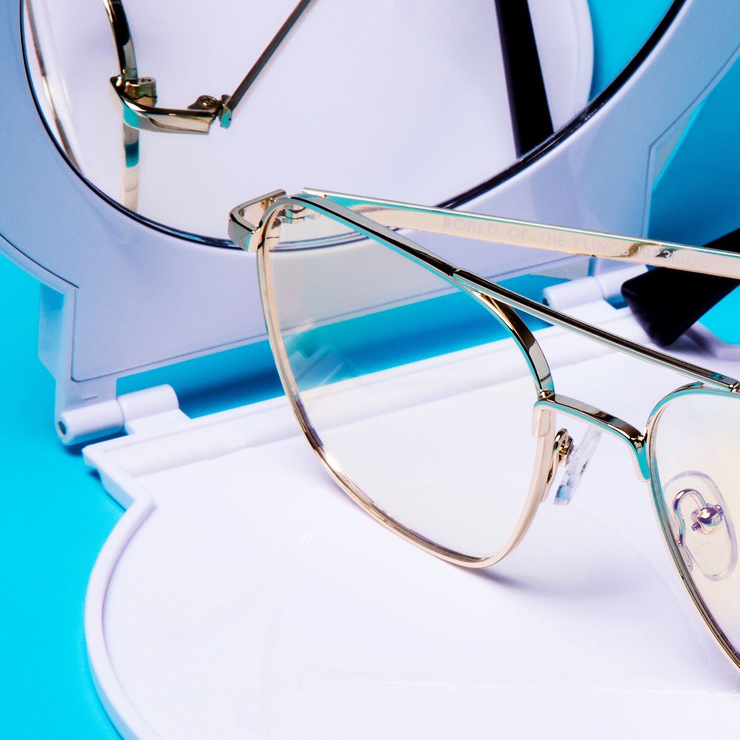 Gold cat-eye metal frame eyeglasses displayed on blue and white background with mirror.