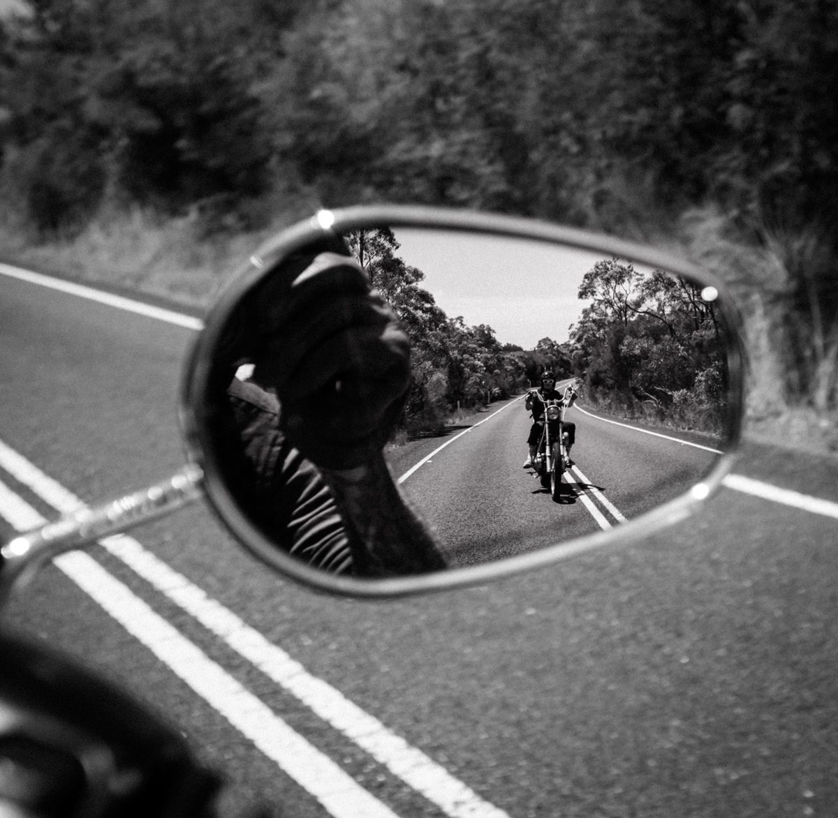 Black and white motorcycle mirror reflection showing a rider on an open tree-lined road.