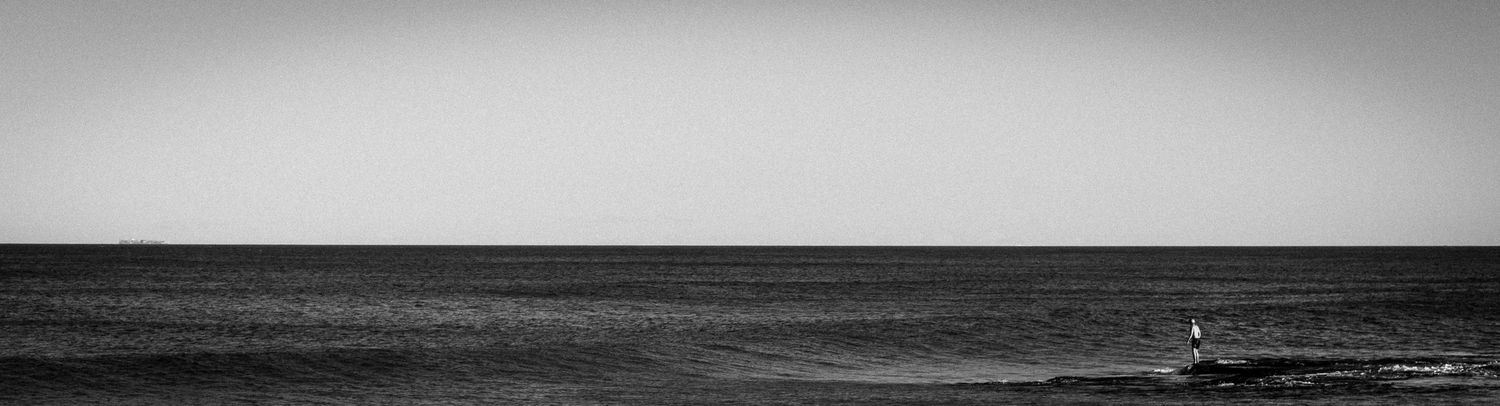 Minimalist black and white panoramic seascape with lone lighthouse on calm ocean horizon.