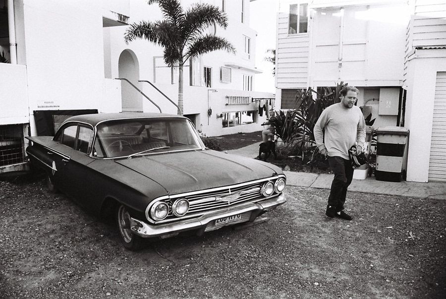 Vintage black and white photo of a man standing beside a classic 1960s Chevrolet in a courtyard.