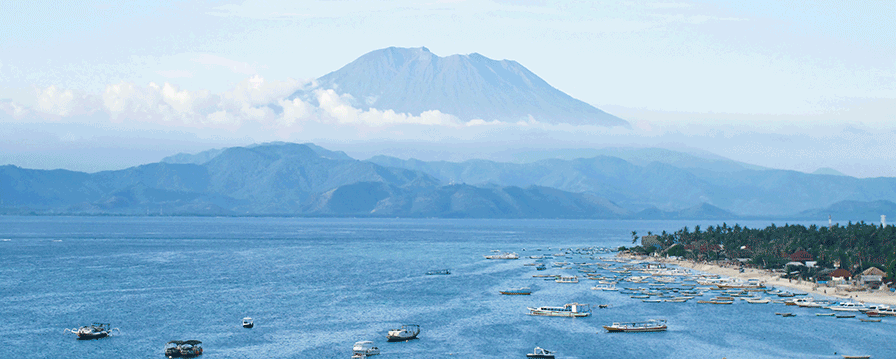 Panoramic view of Bali coastline with fishing boats and volcanic Mount Agung in background.