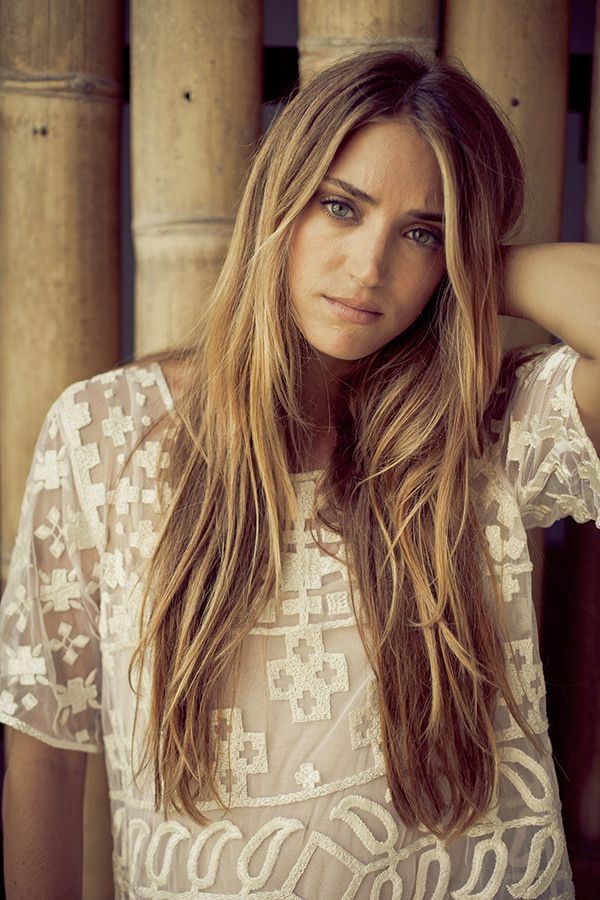 Young woman with long blonde hair wearing white lace top standing near bamboo columns.