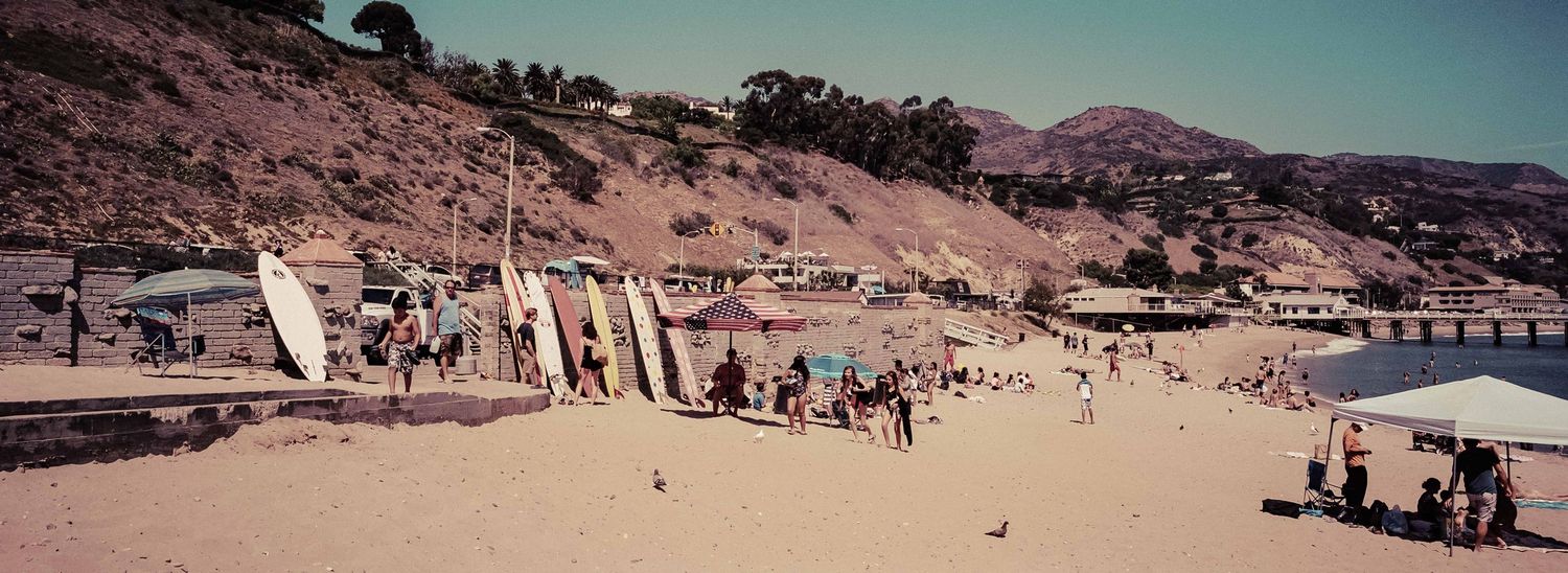 Panoramic beach scene with surfboards lined up and crowds of people near sandy California coastal hillside.