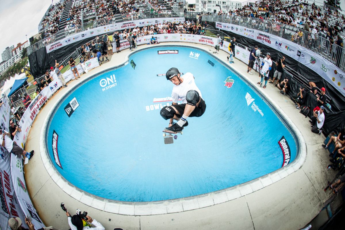 Skateboarder performing aerial trick over blue pool at crowded outdoor skate competition.