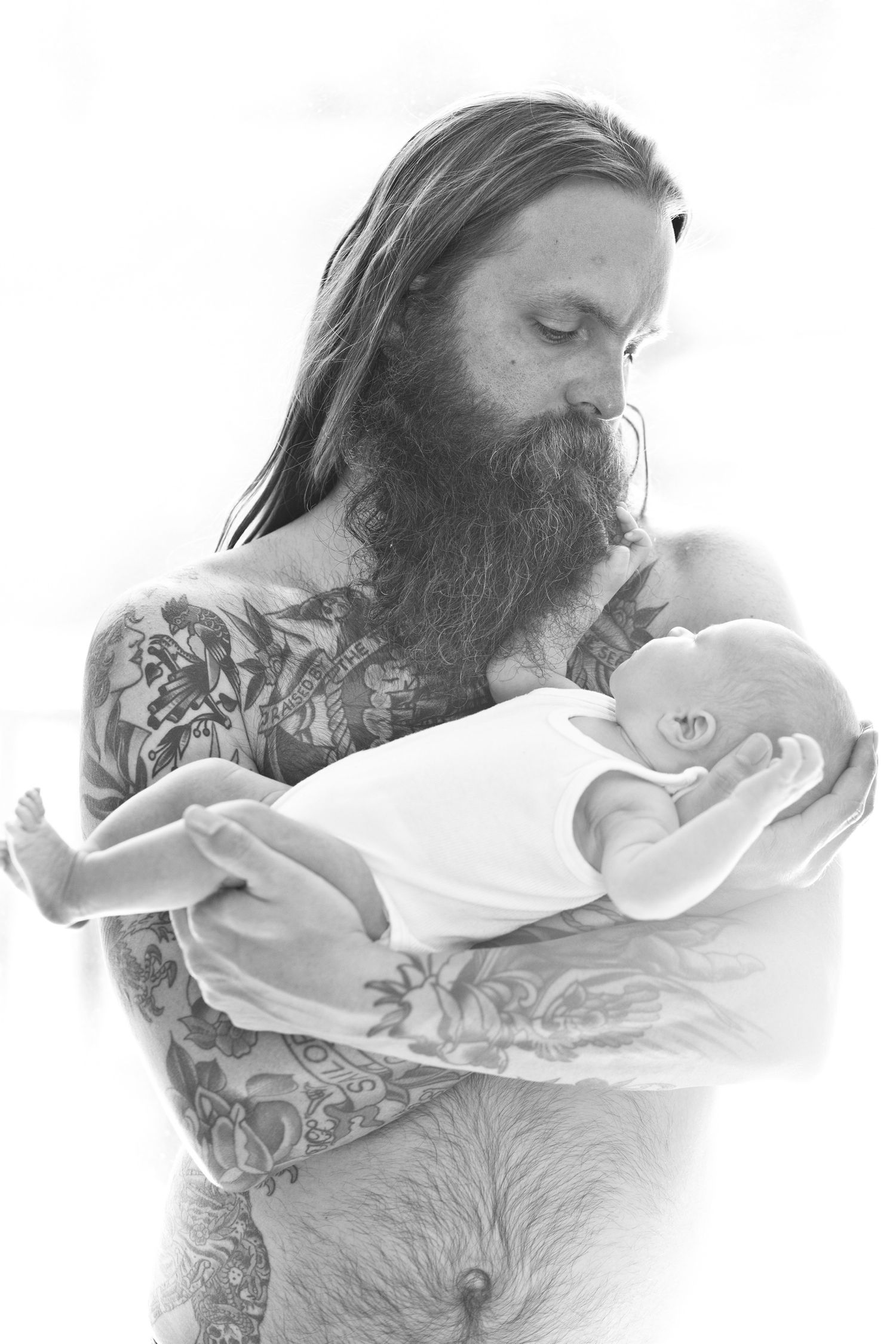 Tattooed man with long hair and beard tenderly holding a newborn baby in black and white.