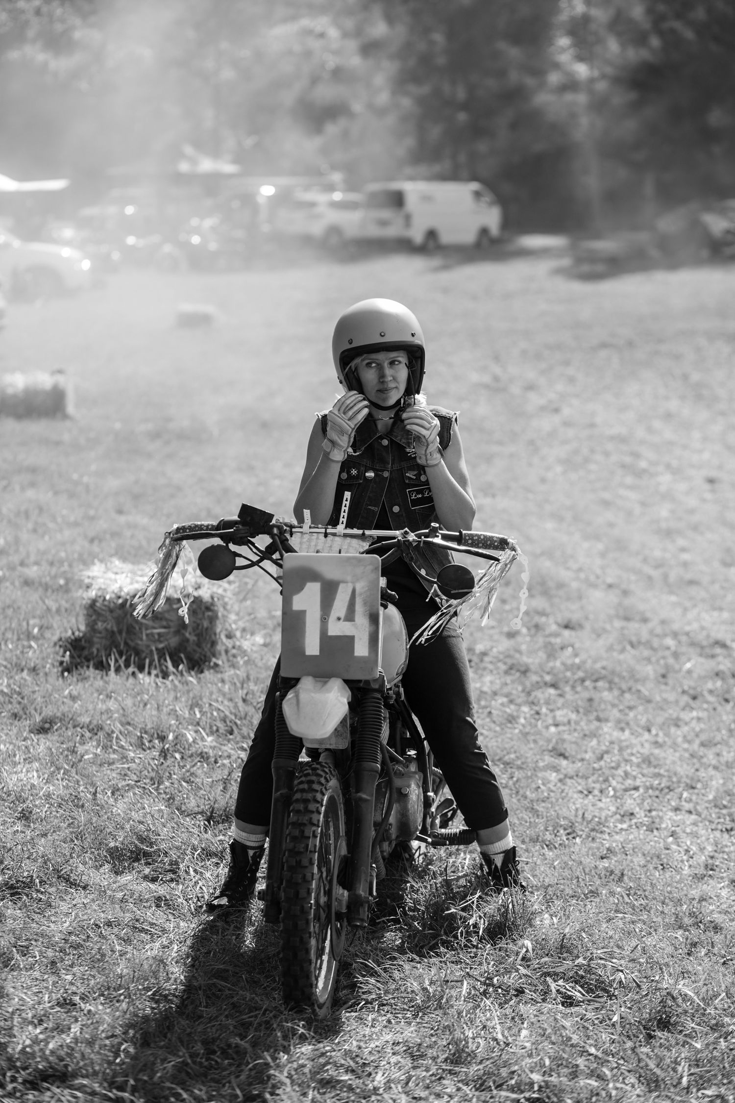 Young motocross rider wearing helmet number 14 sitting on dirt bike in grassy field, black and white photo.