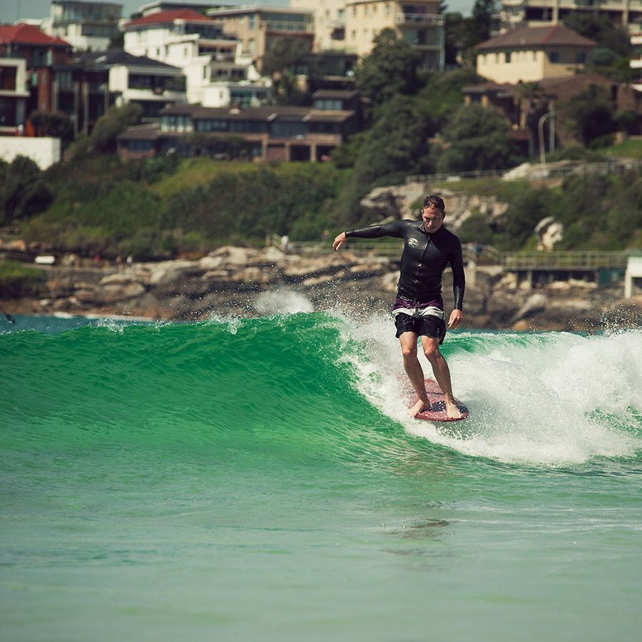 Surfer riding a green wave in a black wetsuit at Bondi Beach with coastal homes in background.