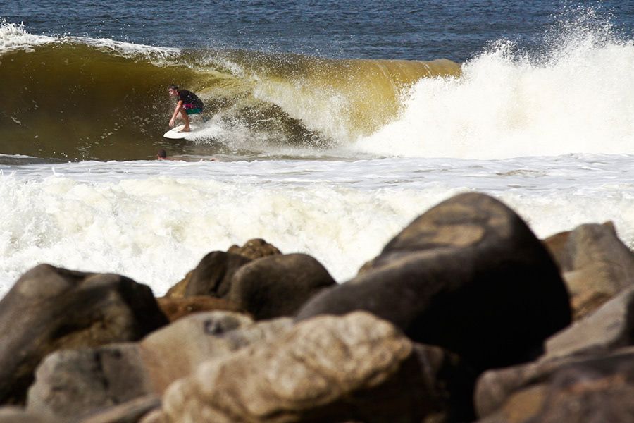 Surfer riding a large wave near a rocky jetty in bright sunny ocean conditions.