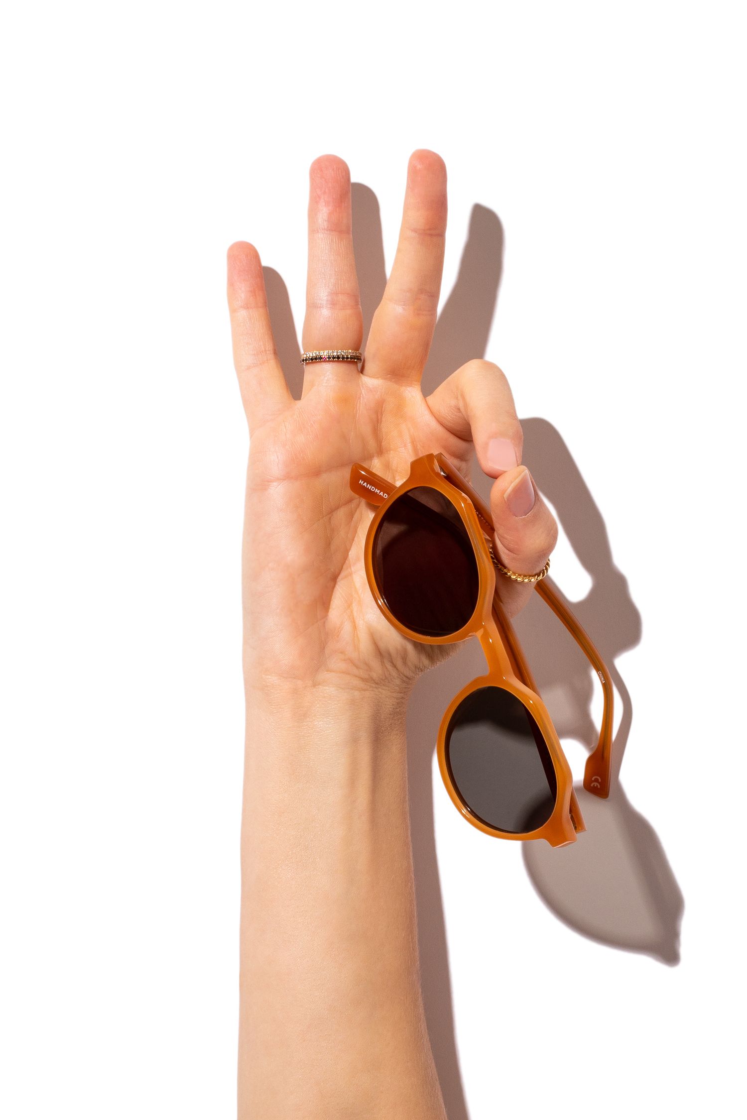 Hand holding amber round sunglasses with dark lenses against a bright white background.