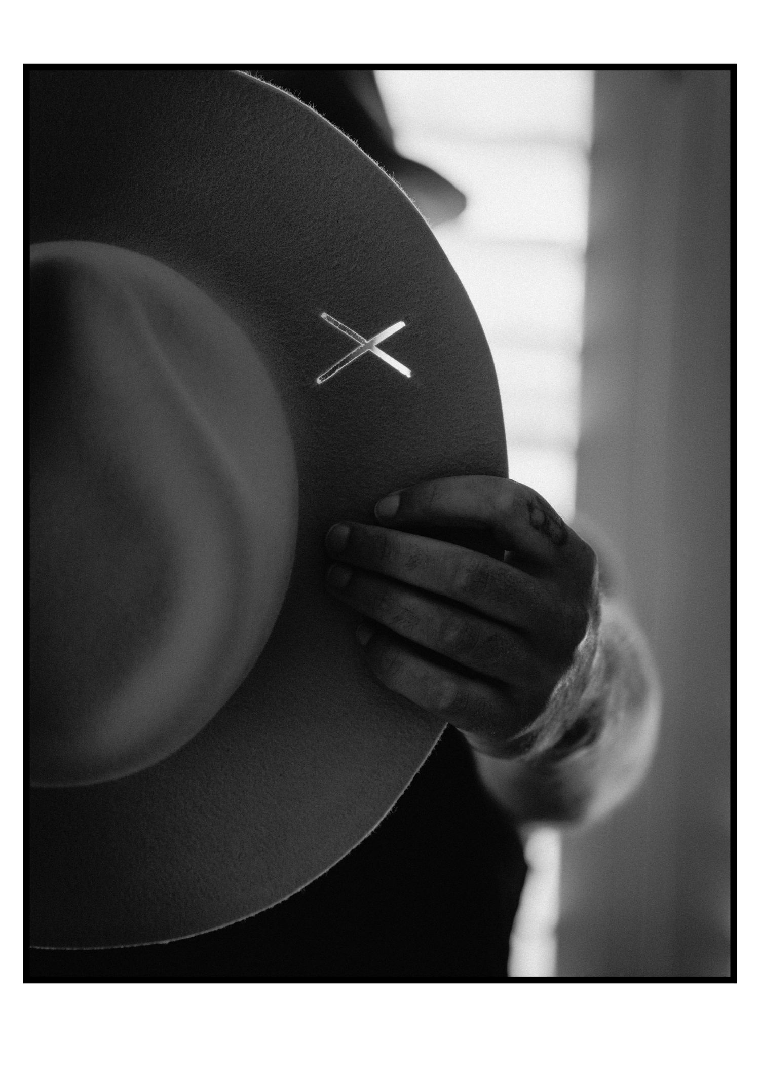 Black wide-brim felt hat with silver cross pin held by hands in shadow.