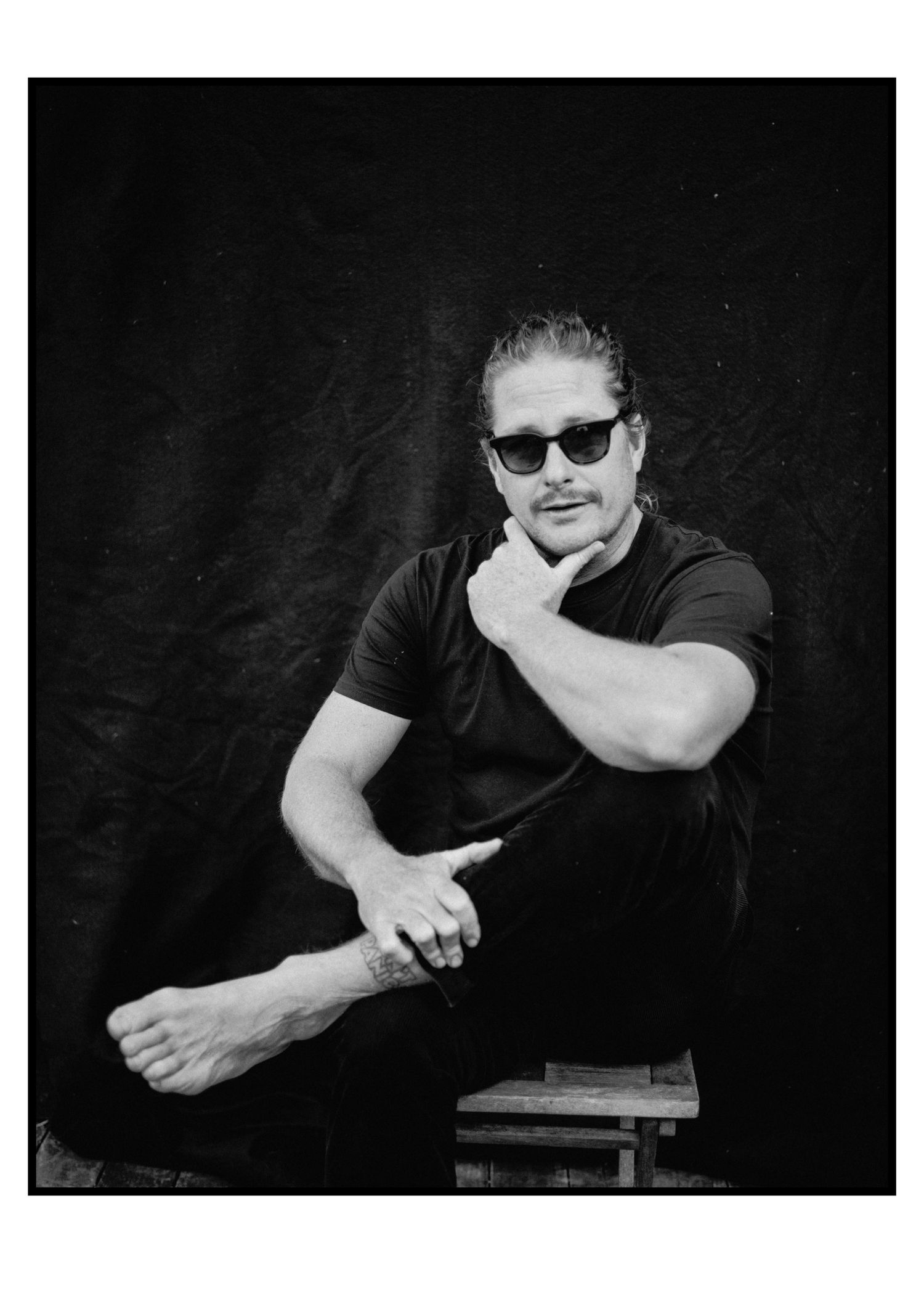 Man in black t-shirt and sunglasses poses thoughtfully on a stool against dark backdrop in black and white portrait.