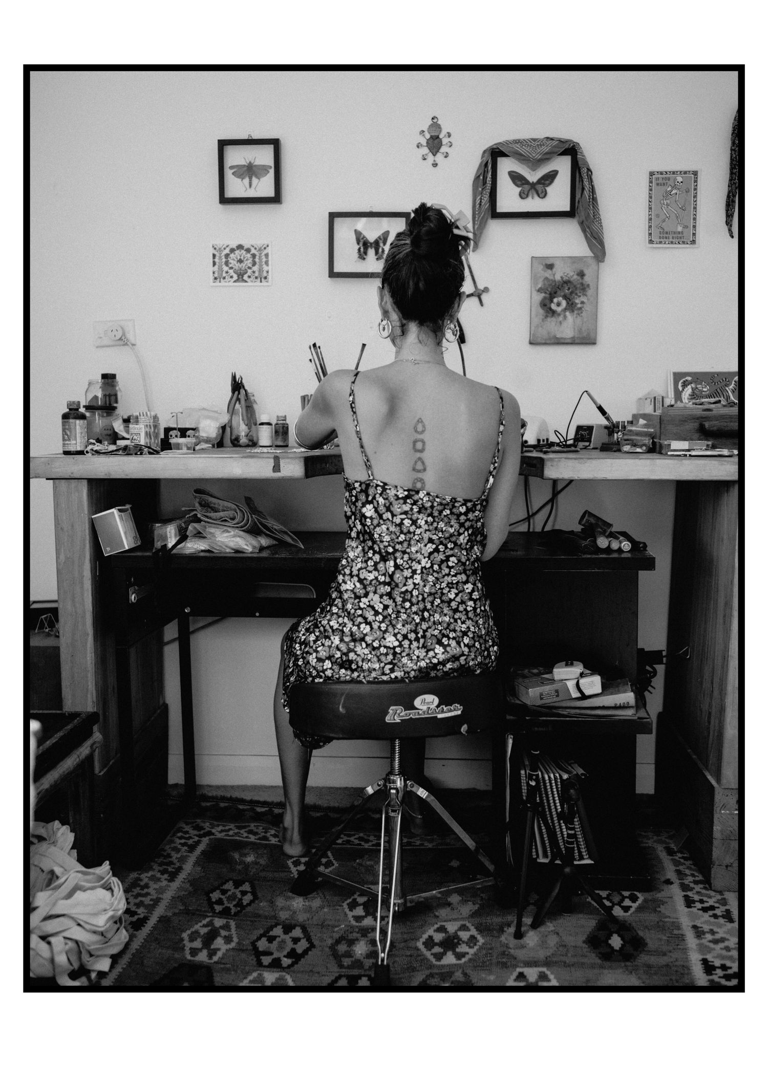 Woman with back tattoos in floral dress sits at cluttered art studio desk in black and white photo.