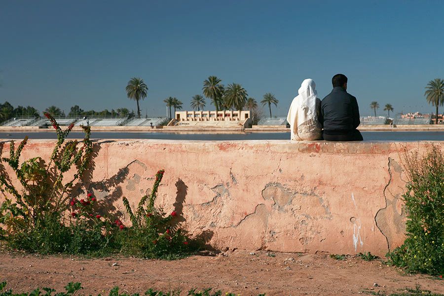 A couple sits together on a weathered pink wall overlooking a sunny North African landscape.