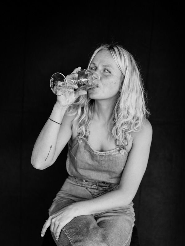 Young blonde woman in denim overalls drinking from a wine glass in black and white portrait.