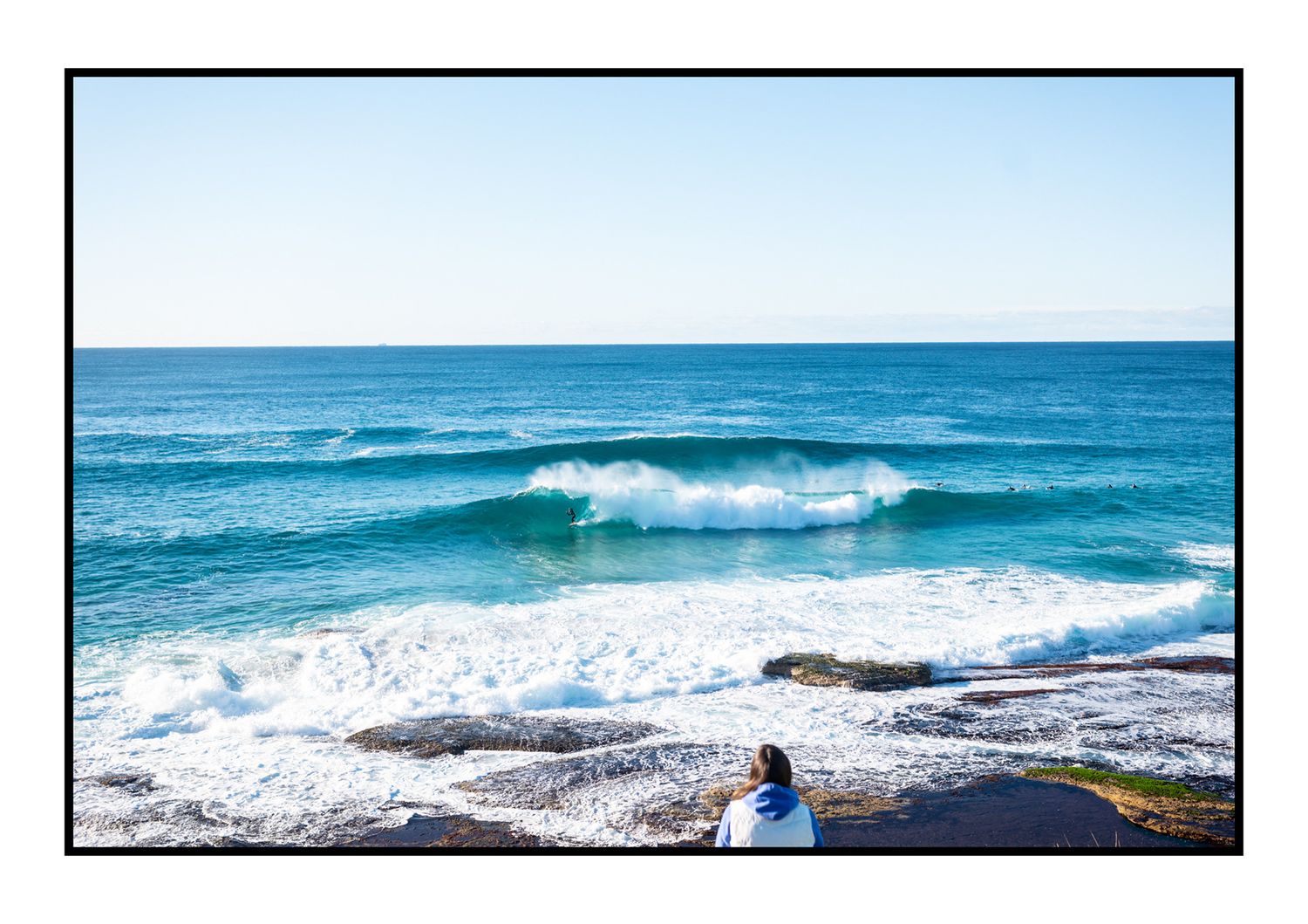 Person sitting on rocks watching large turquoise ocean waves crash on shore.