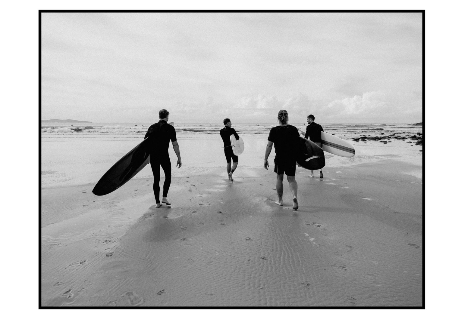 Black and white photo of four surfers carrying boards walking toward ocean waves on sandy beach.