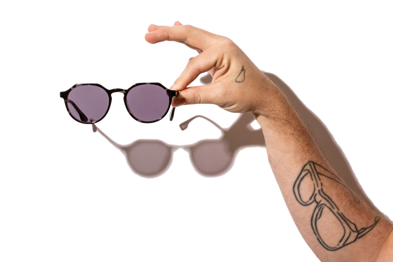 Hand holding tortoiseshell round sunglasses with purple lenses, tattoo visible on forearm.