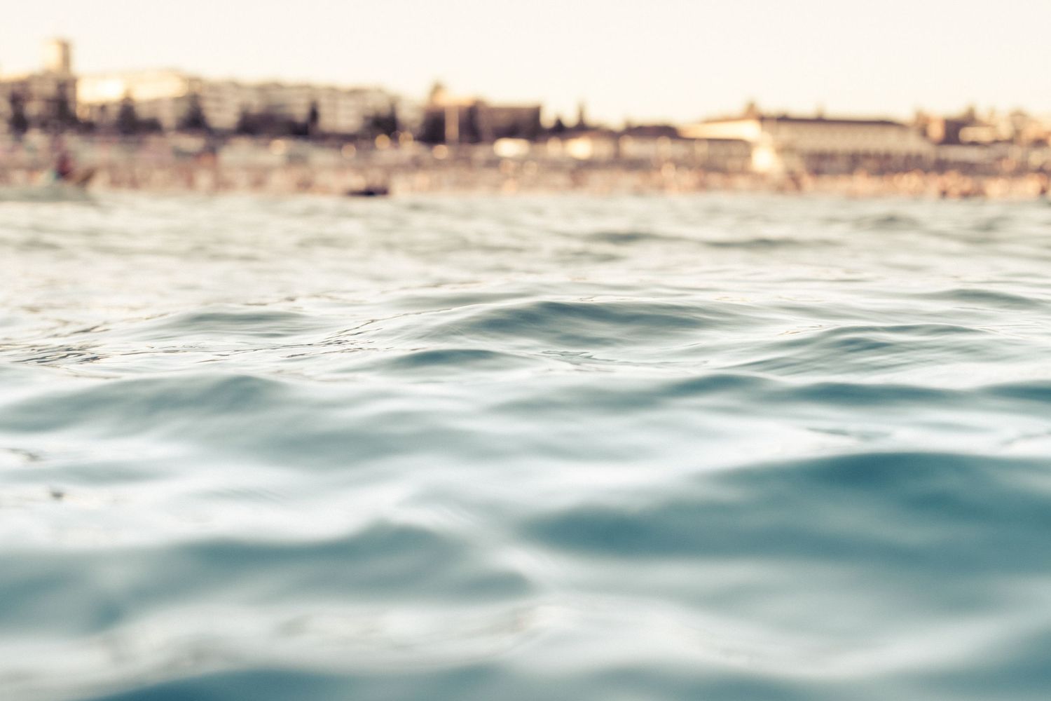 Close-up of calm blue ocean water with blurred coastal buildings in soft bokeh background.