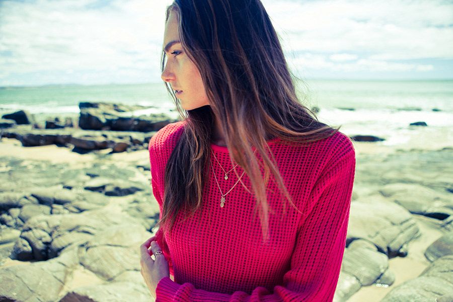 Young woman in red knit sweater standing on rocky beach with ocean in background.