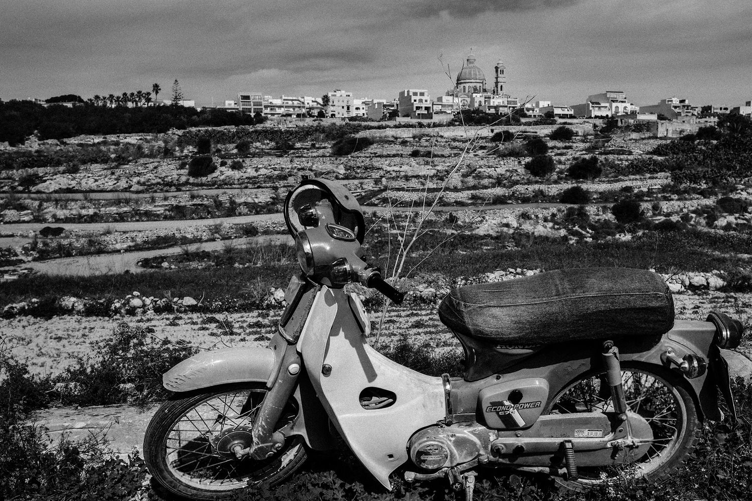 Black and white photo of an abandoned vintage motorcycle in a rocky rural landscape.