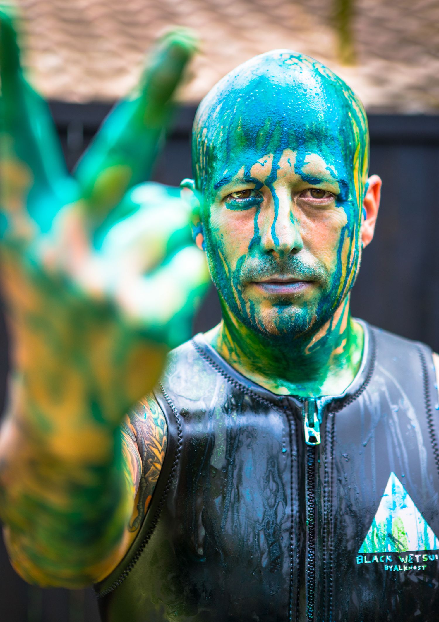 Man covered in green paint wearing a wetsuit making a peace sign gesture outdoors.