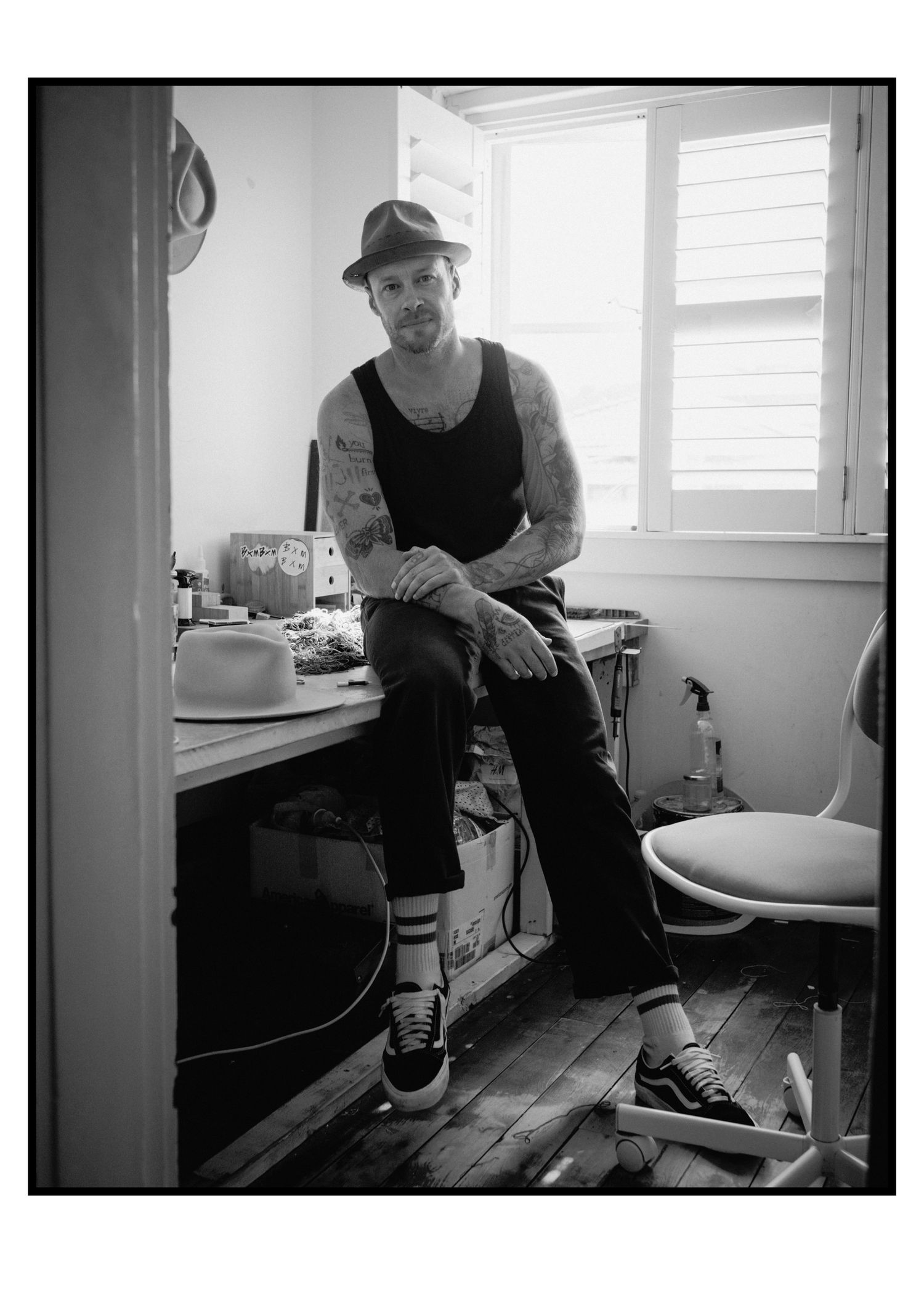 Tattooed artist in fedora hat sitting on desk in creative workspace studio.