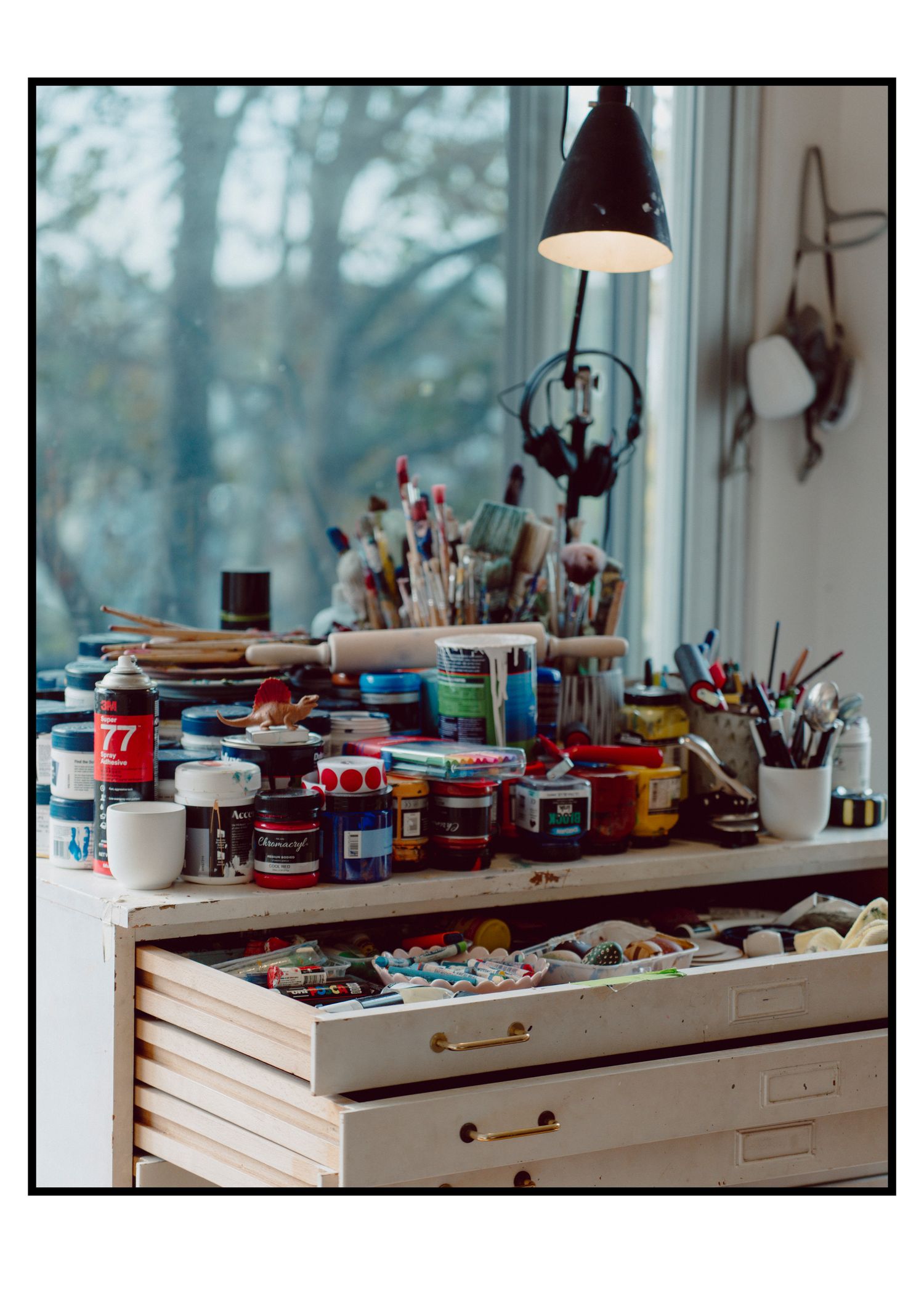 Artist's desk with paint jars, brushes, and supplies near a window with lamp.