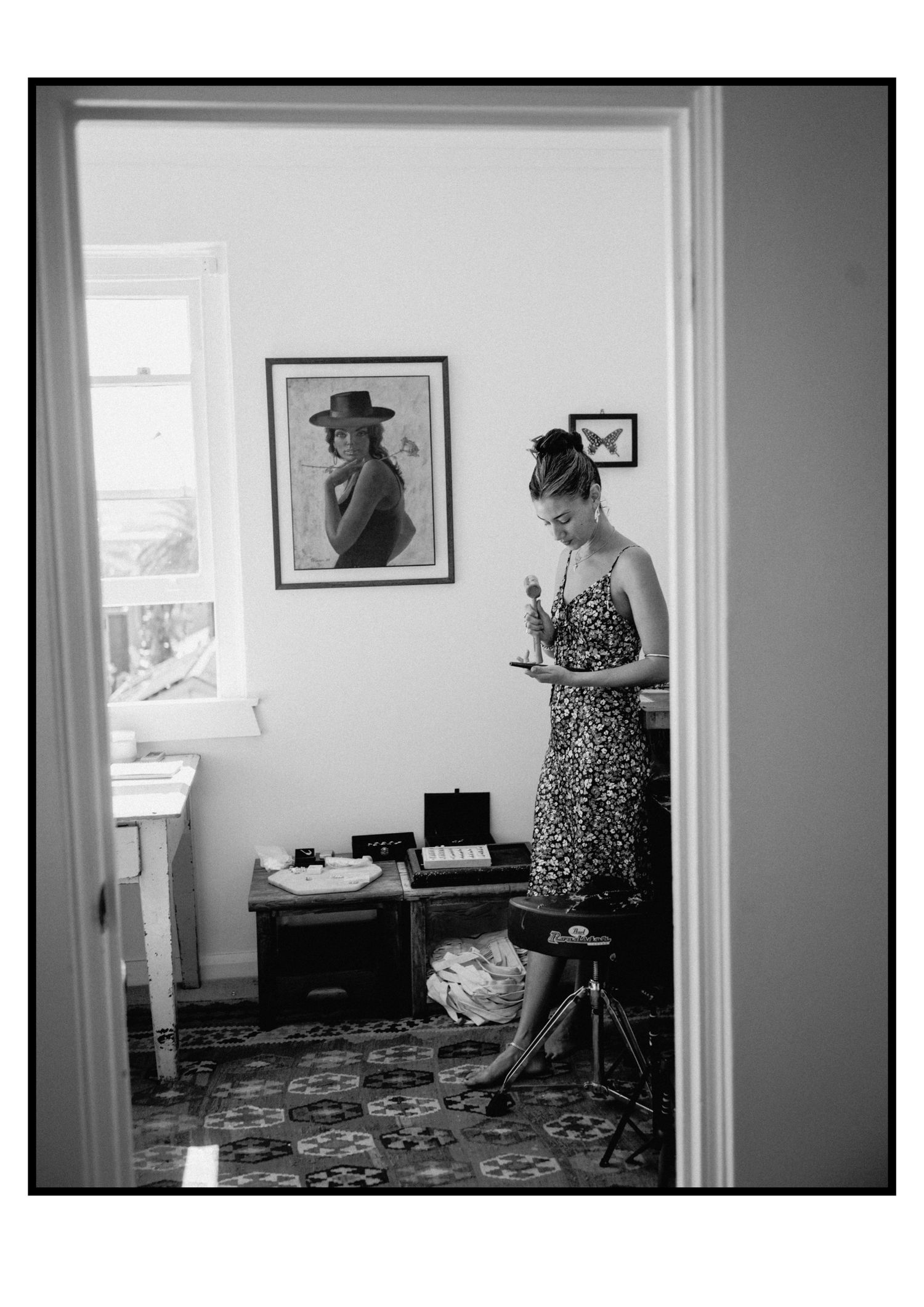 Woman in floral dress examines small object in artistic studio room viewed through doorway in black and white.