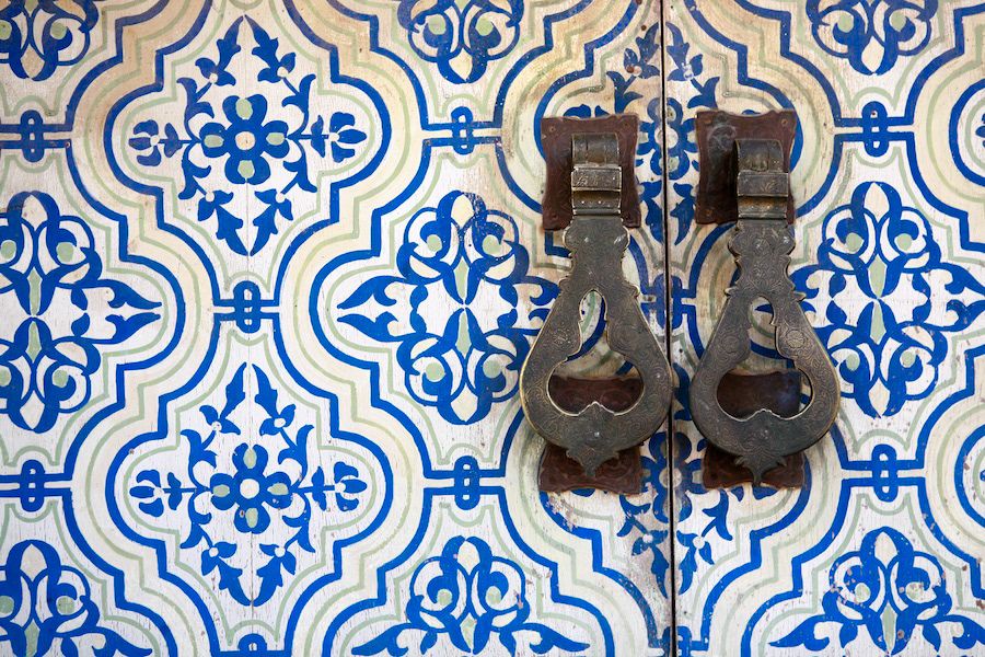 Ornate blue and white Moroccan tilework with two vintage iron door knockers on a decorative entrance.