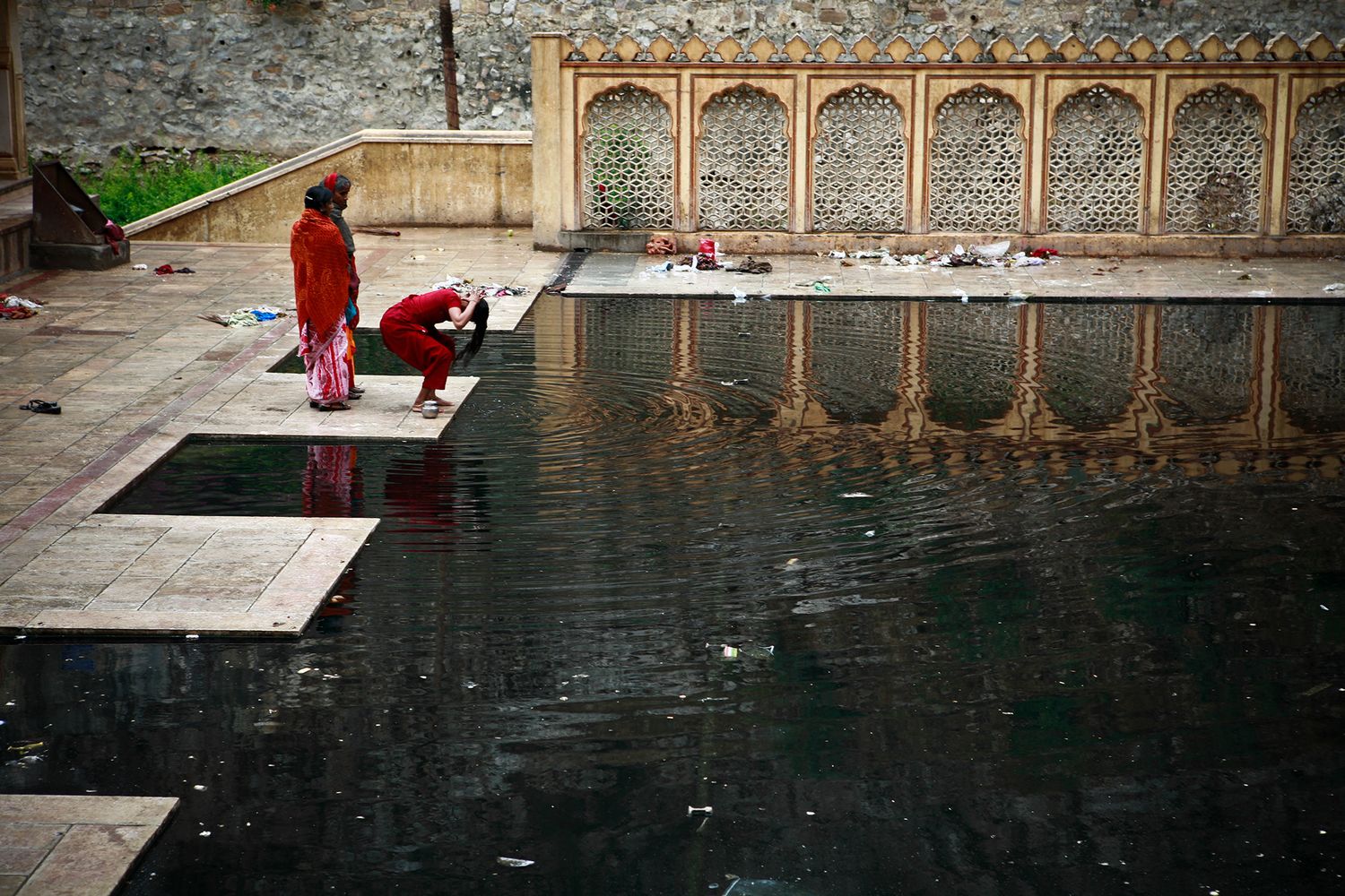 Two women in traditional dress perform ritual washing beside a dark sacred temple pool in India.