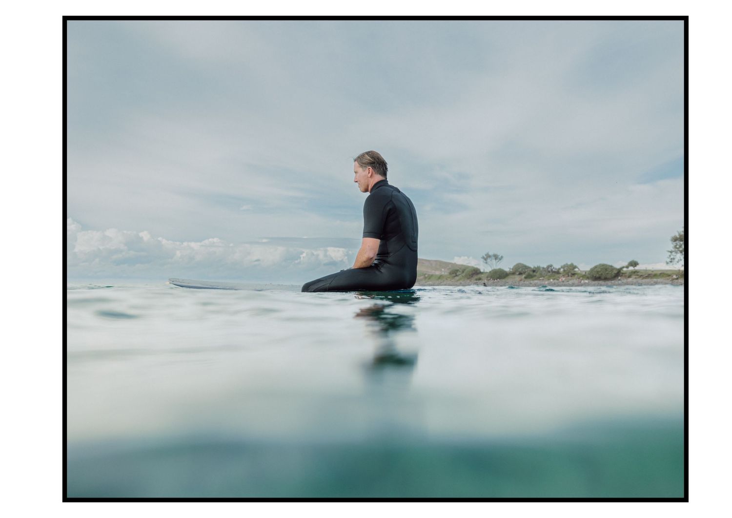 Surfer in wetsuit sitting on surfboard in calm ocean waters with cloudy sky.