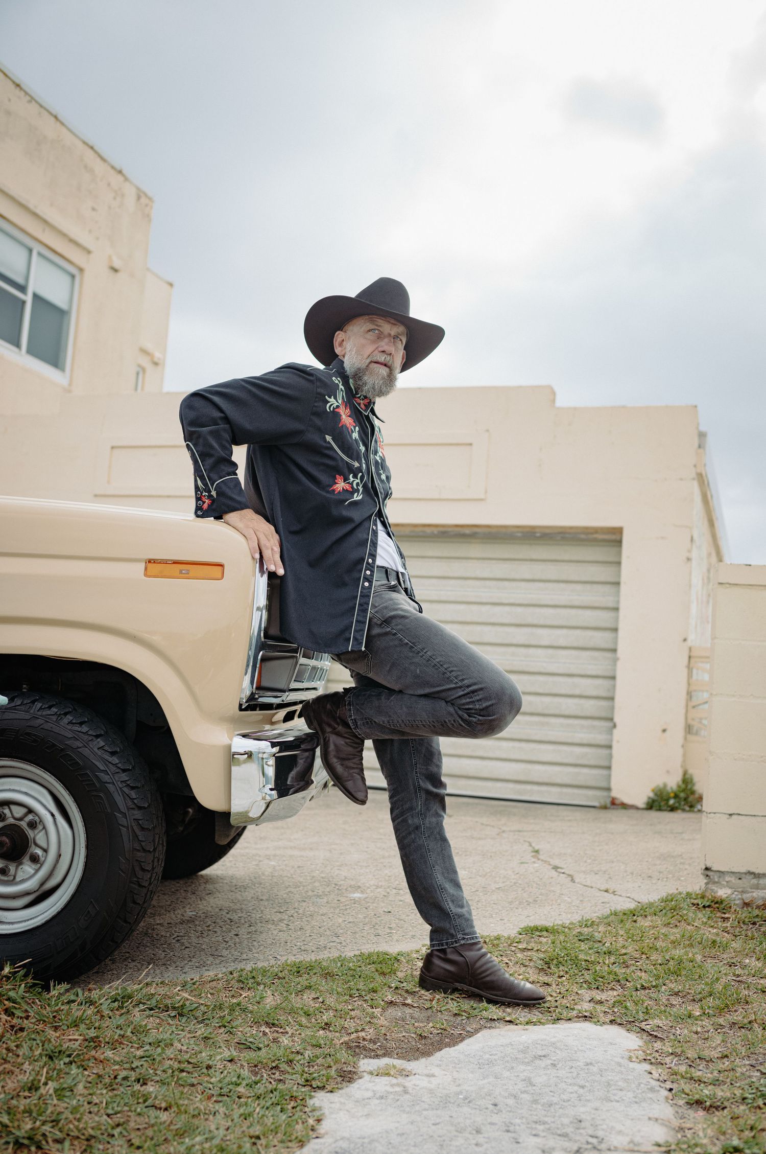 Man in black cowboy hat and embroidered western shirt posing with foot on vintage cream pickup truck outdoors.