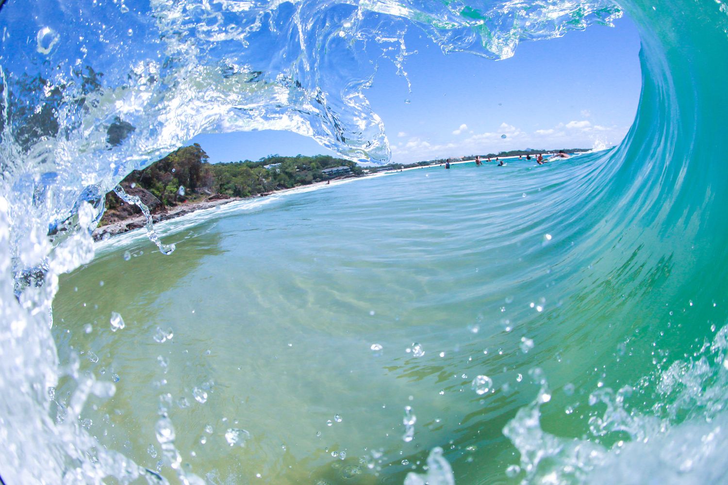 A stunning turquoise wave curls over a sandy beach with a rocky headland in the background.