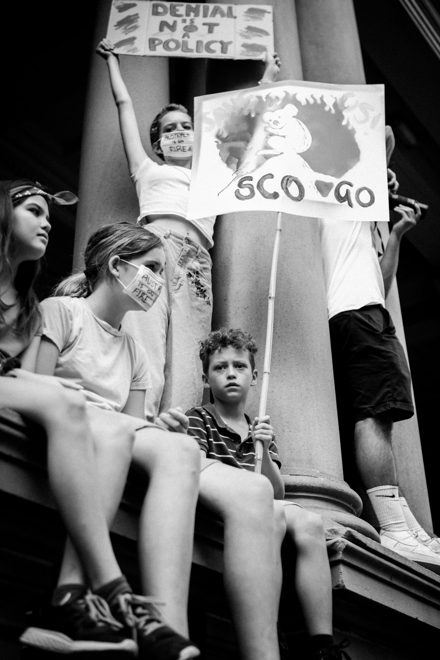 Black and white photo of children at a climate protest holding signs reading Denial Not Policy and Sco Go.