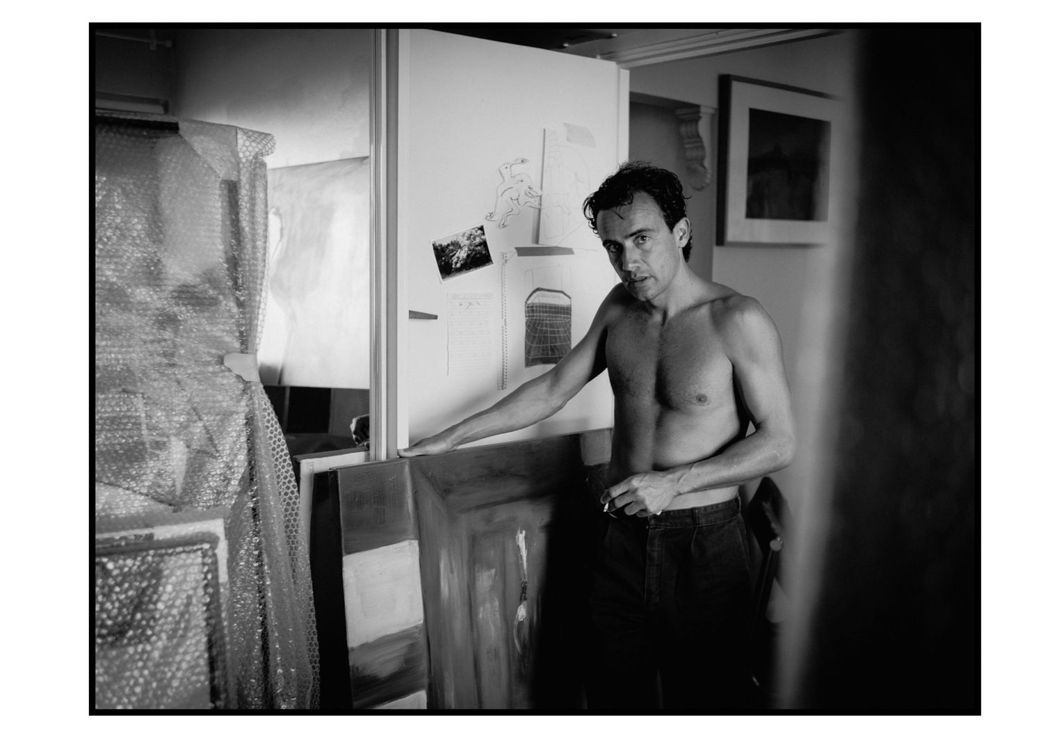 Shirtless man stands beside canvases in art studio in black and white documentary photograph.