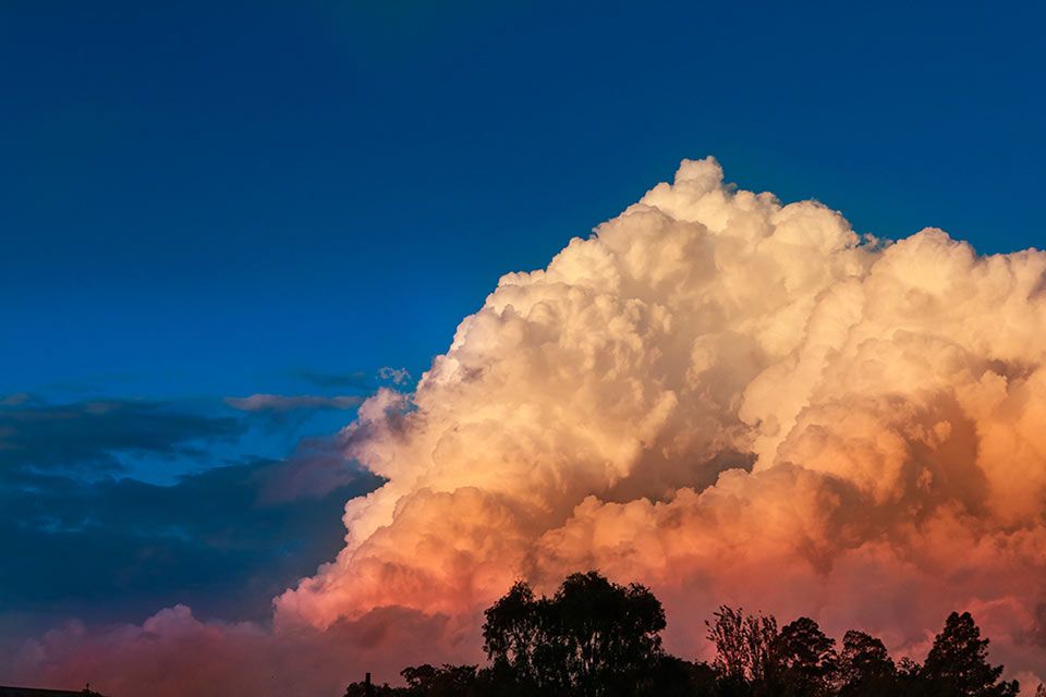 Dramatic cumulus storm cloud illuminated in orange and pink hues against a deep blue evening sky.