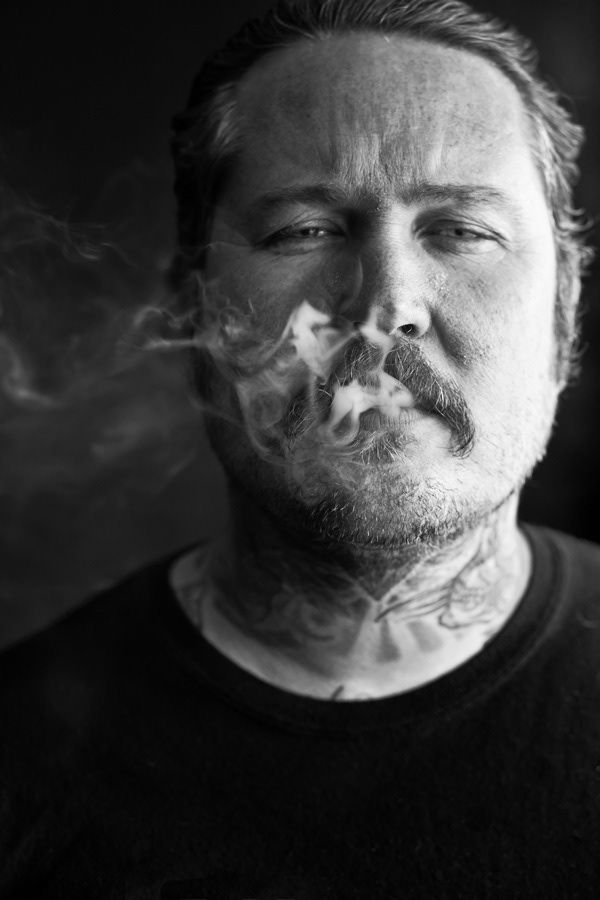 Black and white portrait of bearded man with neck tattoos exhaling smoke.